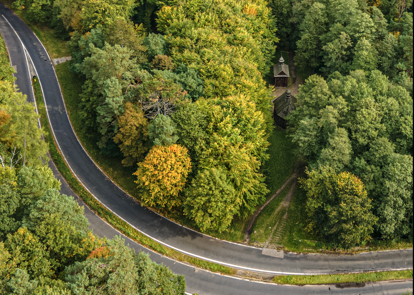 Las Łagiewnicki - zdjęcie z lotu ptaka przebarwiających się koron drzew. W dole kadru zakręt drogi, w centrum - kapliczki w Lesie Łagiewnickim
